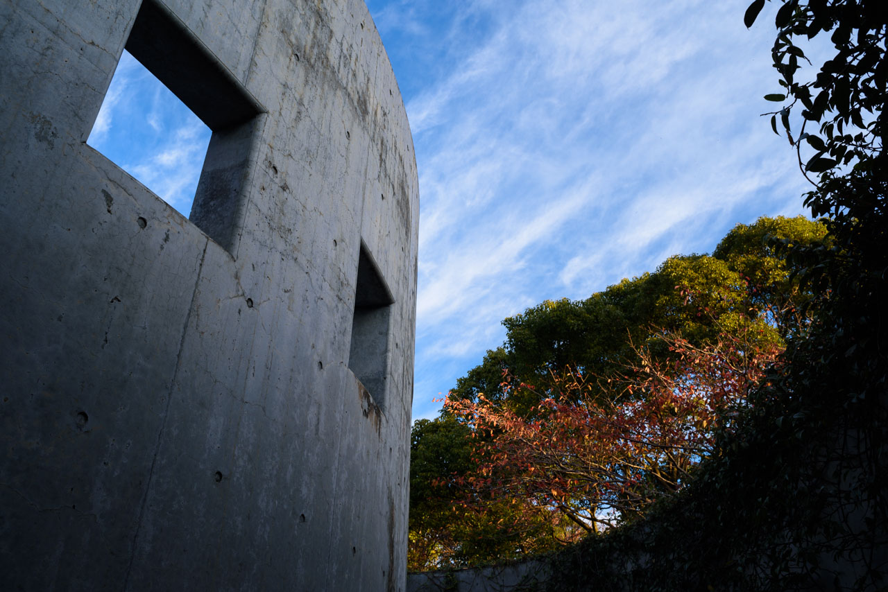 Looking up at the outer wall of the museum, the blue sky, and the trees in the park. The blue sky can be seen beyond the square hole in the outer wall.