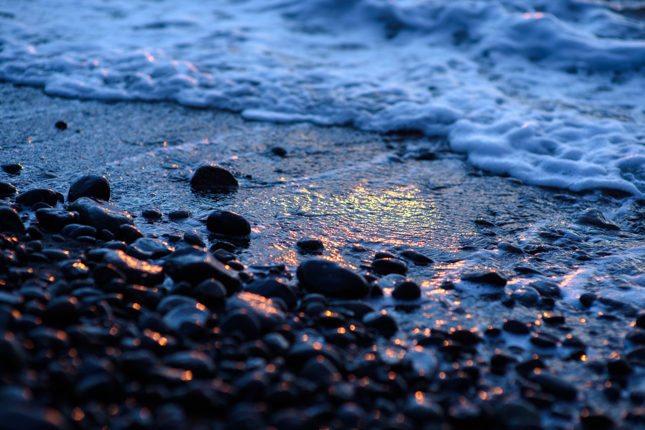 波打ち際の砂浜と丸石に朝日のオレンジ色がきらめく The orange of the morning sun sparkles on the sand and cobble stones at the edge of the waves. 