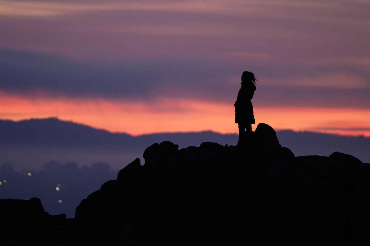 Silhouette of a woman standing on rocky shore at Lovers Point Park in Monterey during dawn glow